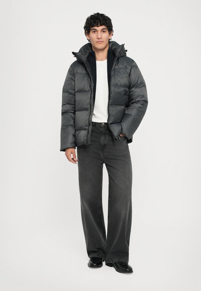 Dunkelgraue Pufferjacke mit hohem Kragen, Reißverschluss und kastigem Schnitt, getragen über einem weißen Shirt und schwarzen weiten Hosen.