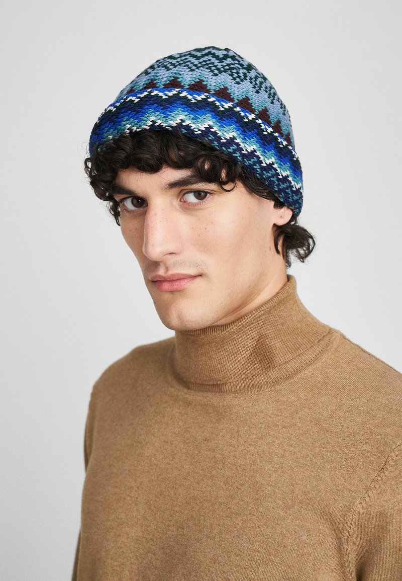 Knit beanie in blue tones with geometric patterns and a textured finish. Worn with a brown turtleneck sweater.