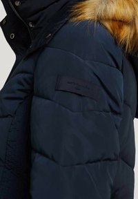 Granatowa kurtka puchowa z kapturem wyściełanym futrem, z pikowaną fakturą, zatrzaskami i naszywką z logo na górnej części rękawa.