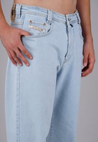 Mann in hellblauen Jeans mit der Hand im vorderen Taschen gegen einen neutralen grauen Hintergrund.