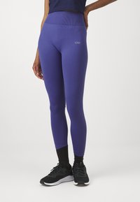 Lilla leggings med høy midje, laget av elastisk materiale. Har en logo på venstre side og en jevn tekstur. Svart treningssko er brukt.