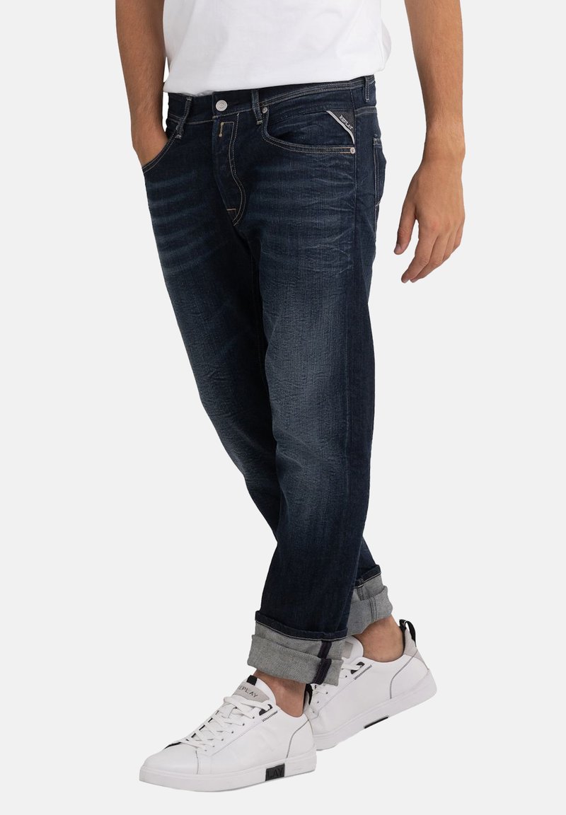 Mann in dunkelblauen, hochgekrempelten Jeans und weißen Sneakers, der mit einer Hand in der Tasche vor einem schlichten Hintergrund steht.