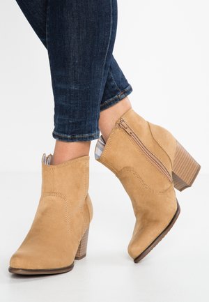 Personne portant des bottines en daim marron clair avec talons blocs et fermetures éclair latérales, associées à un jean skinny bleu foncé sur fond blanc.