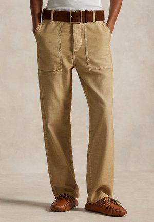 Άτομο που φοράει μπεζ παντελόνια φαρδιά με μεγάλες μπροστινές τσέπες, καφέ ζώνη από σουέτ και καφέ δερμάτινα μοκασίνια, στέκεται με τα χέρια στις τσέπες.