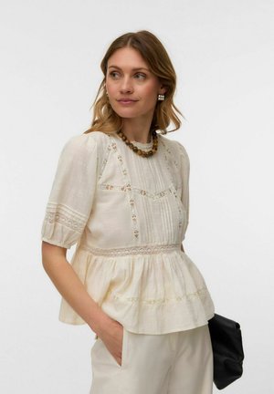 Kvinne iført kremfarget bluse med blonderkant og pufferm, og perlekjede, holder en svart clutch, ser til siden.