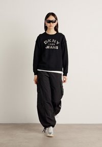 Svart hettegenser med "DKNY 1989 JEANS" grafikk, kombinert med løse svarte cargobukser. Sølv sneakers fullfører antrekket. Tilbehøret inkluderer solbriller.