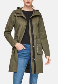 Giacca verde oliva con cappuccio, chiusura a zip, due tasche laterali e vita regolabile con cordino. Tessuto liscio e leggero.