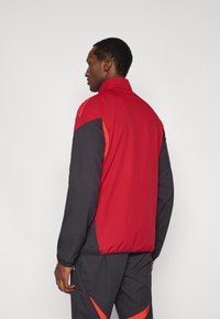 Red and black athletic jacket with a high collar, elastic cuffs, and contrasting side panels. Smooth fabric with a fitted design.