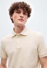 Νεαρός άνδρας με καστανά σγουρά μαλλιά φοράει ένα υφασμάτινο μπεζ κοντομάνικο πόλο πουκάμισο, κοιτάζοντας ελαφρώς προς τα πάνω με φόντο απλό λευκό.