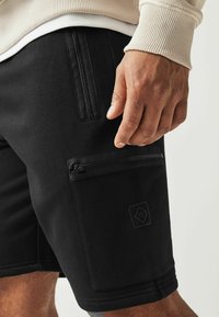Shorts noirs en tissu lisse, dotés de deux poches zippées et d'un logo discret. L'ourlet est fini par un bord net.