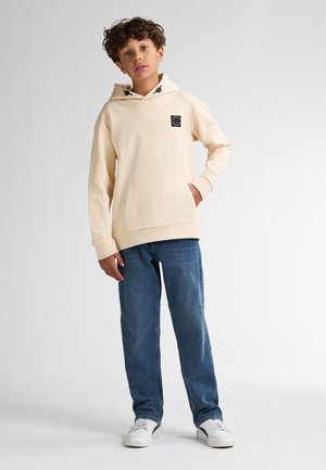 Beige hoodie met een voorzak en zwart logo, gecombineerd met blauwe denim jeans en witte sneakers. Effen achtergrond.