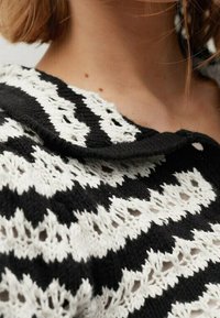 Gros plan sur un pull tricoté noir et blanc avec un motif en zigzag texturé et un col large replié porté par une personne.