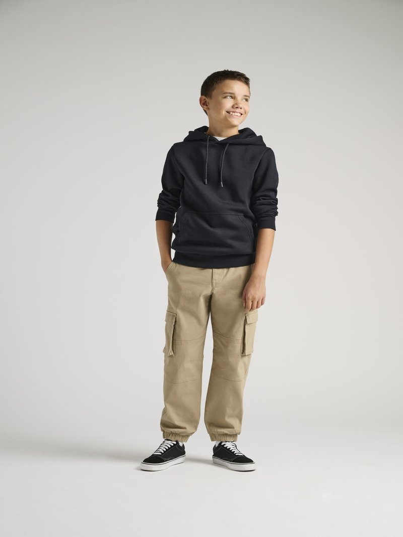 Svart hettegenser med frontlomme, beige cargo-bukser med sidelommer, og svarte sneakers med hvite detaljer, stående på en ensfarget bakgrunn.