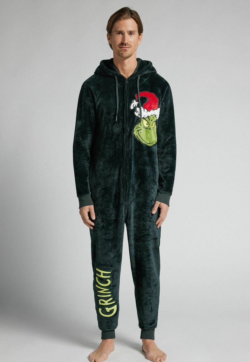 Zielony, pluszowy kombinezon z wizerunkiem twarzy Grincha i czapką Mikołaja na piersi, z napisem "GRINCH" w pogrubionym żółtym kolorze na lewej nodze.