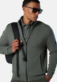 Maglione di colore verde scuro, realizzato in tessuto a maglia testurizzato. Presenta una cerniera frontale completa, tasche laterali e orlo a coste. Supportato da una cintura nera per lo zaino.