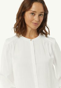 Biała bluzka z długim rękawem, z delikatnymi fałdami na ramionach oraz okrągłym dekoltem z małym zapięciem na guzik. Gładka tekstura materiału.