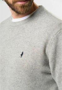 Hombre vistiendo un suéter de punto gris claro con una pequeña figura bordada en azul marino oscuro en el lado izquierdo del pecho, camisa blanca debajo.