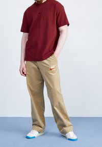 Bordová bavlněná tričko s malým logem Nike; béžové kalhoty s logem "Nike"; bílé boty s modrými akcenty; jednobarevné pozadí.