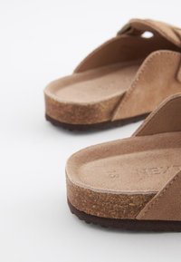 Brūnas zamšādas iešļūcenes ar teksturētu korķa zoli, kurām ir plata pirksta siksna un noapaļota pēdas forma ērtībai.