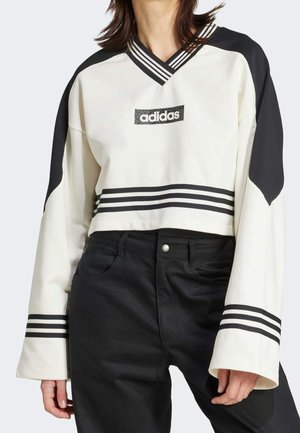 Henkilö pukeutunut valkoiseen, lyhytmalliseen Adidas-collegepaitaan, jossa on mustat raidat hihoissa, helman ja rinnassa olevan logon ympärillä, yhdistettynä korkeavyötäröisiin mustiin housuihin.