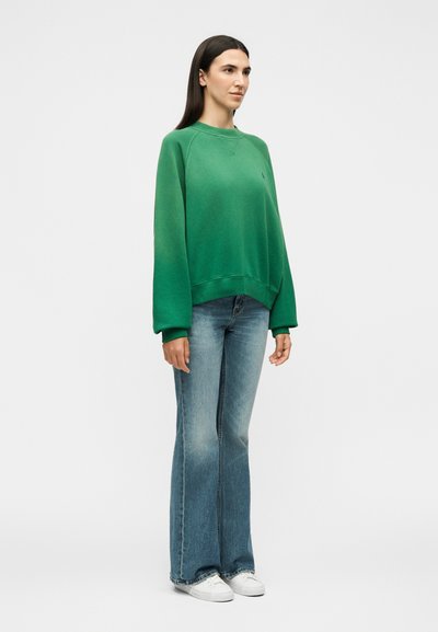 Γυναίκα που στέκεται φορώντας μια πράσινη μεγάλη φαρδιά μπλούζα, ξεθωριασμένο μπλε τζιν bootcut και λευκά αθλητικά, σε απλό ανοιχτό φόντο.