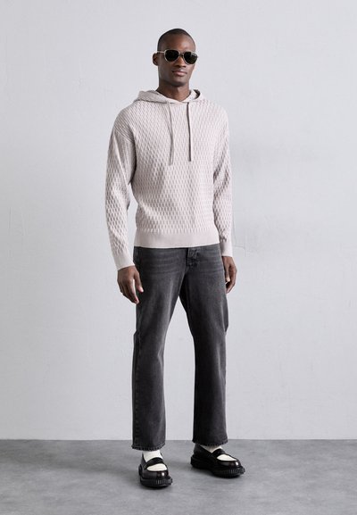 Haut de survêtement texturé gris clair avec un cordon de serrage, associé à un jean gris foncé et des chaussures noires massives. Le mannequin porte des lunettes de soleil rondes.