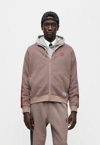 Κόκκινο και καφέ καρό σακάκι με φερμουάρ, με ανοιχτό γκρι φούτερ με κουκούλα από κάτω, σε συνδυασμό με παντελόνι που ταιριάζει και ριγέ καπέλο.