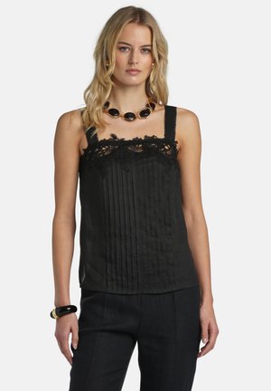 Szőke nő fekete ujjatlan felsőben, csipkedíszítéssel és pliszírozott részletekkel, fekete nadrágban, vastag fekete nyaklánccal és fekete karkötővel.