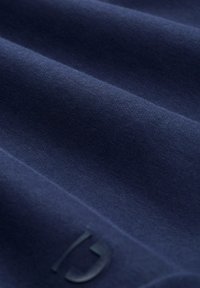 Mornarijski materijal s glatkom teksturom, koji sadrži suptilne nabore. Mali, tamni logo vidljiv je u donjem kutu, što poboljšava dizajn.