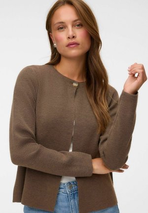 Jeune femme aux longs cheveux châtain clair portant un cardigan marron boutonné, un haut blanc et un jean bleu, posant avec un bras plié.