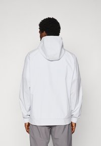 Homme aux cheveux bouclés noirs portant une veste à capuche blanche et un pantalon gris, se tenant de dos devant un fond uni.