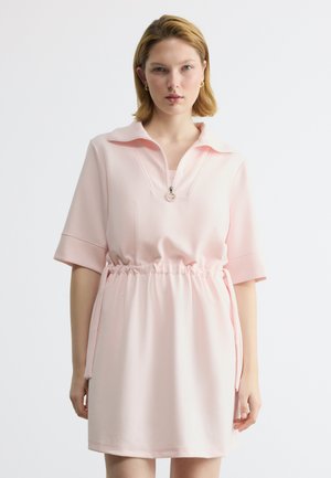 Γυναίκα που φοράει ανοιχτό ροζ κοντομάνικο φόρεμα με φερμουάρ στο γιακά και σούρες στη μέση, στέκεται μπροστά σε απλό λευκό φόντο.