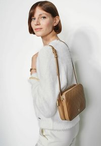 Karmelowa torba z pikowanej tkaniny z odpinanym paskiem, prostokątna forma, złote akcenty, noszona na ramieniu na białym tle.