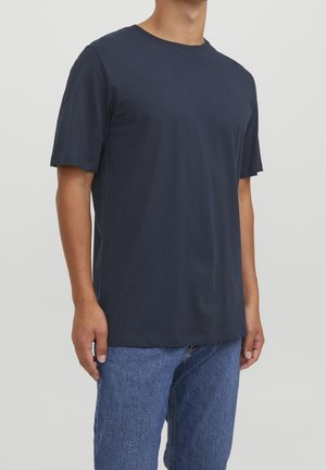 Homme portant un t-shirt bleu marine uni à manches courtes et un jean bleu sur un fond clair uni.