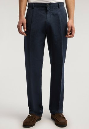 Mann trägt marineblaue gerade geschnittene Hosen und braune Wildleder-Loafers, steht mit entspannt an den Seiten hängenden Armen.