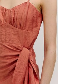 Roze gekleurde jurk met een geplooide lijfstijl, verstelbare spaghettibandjes en een gewikkelde zijkant met een gestrikt knoopje. Gestructureerde kreukstof.