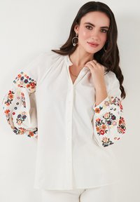 Witte blouse met lange, opgeblazen mouwen en kleurrijke bloemenborduursels. Zachte stof met een geraffineerde halslijn.