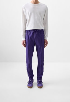 Osoba ubrana w białą koszulę z długim rękawem, fioletowe spodnie dresowe z bocznymi paskami oraz pasujące fioletowe trampki, stojąca na jednolitym tle.