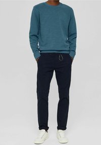Persona con un suéter azul de cuello redondo, pantalones oscuros navy con un clip de metal, manos en los bolsillos y zapatillas blancas sobre un fondo gris.