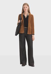 Brauner Cordjacke, schwarzer Pullover, gemusterter Kragen, weite dunkle Jeans und schwarze Tragetasche. Braune Schuhe runden das Outfit ab.