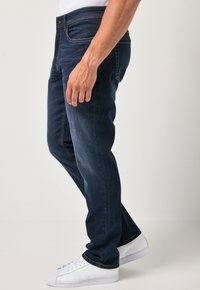 Jeans de mezclilla azul oscuro con corte recto, que presentan costuras amarillas, bolsillos frontales y cierre de botón. Combinados con zapatillas blancas.