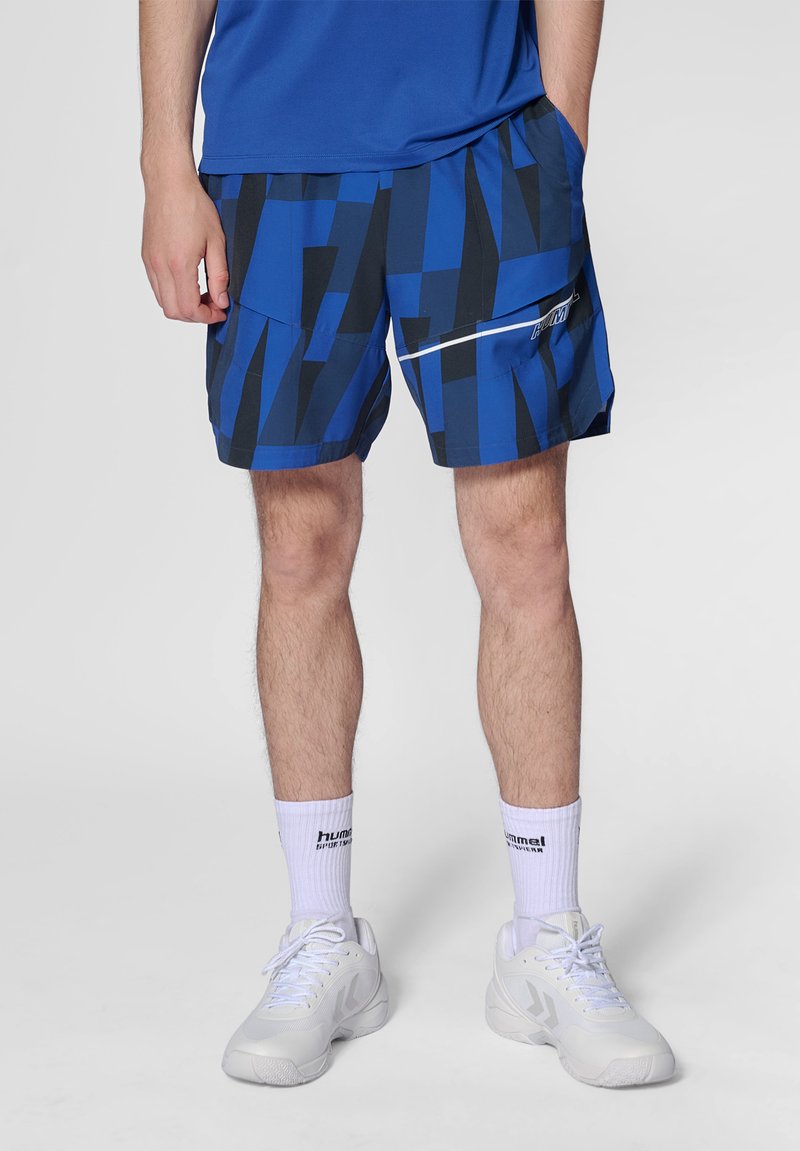 Man draagt blauw-zwart patroon sportshorts, witte Hummel sportsokken, witte sneakers, staand met één hand in de zak.