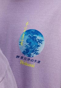 Lavendelfärgad bomullströja med en blå cirkulär grafik som har blommönster och text i gult som framhäver "Hokusai".