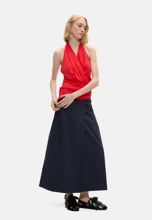 Kobieta w czerwonym topie bez rękawów na szyi halter, długiej granatowej spódnicy i czarnych sandałach z frędzlami, stoi ze splecionymi rękami na wysokości talii.
