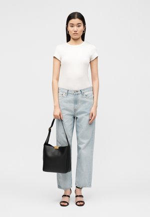 Mujer de pie mirando hacia adelante, lleva una camiseta blanca de manga corta, vaqueros azul claro, sandalias negras y sostiene un bolso negro a su lado.