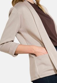 Beige lehký blazer s dlouhými rukávy a ohrnutými manžetami; má přední kapsu a otevřený střih. Nositeli ho mají na hnědém topu.