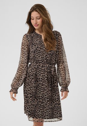 Femme portant une robe léopard mi-longue à manches longues et ceinture à la taille, debout devant un fond uni gris clair.