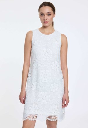 Frau in ärmellosem weißem Spitzenkleid mit großen Blumenmustern und gezacktem Saum, steht vor hellem, einfarbigem Hintergrund.