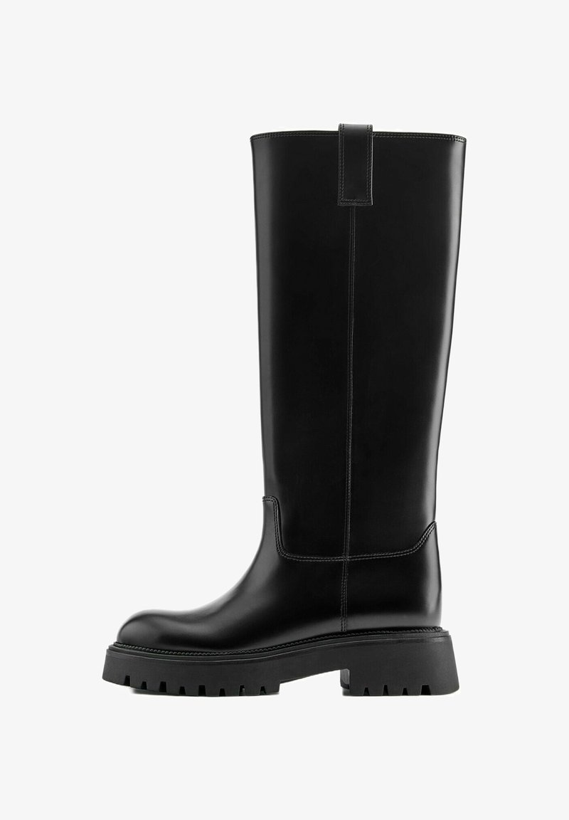 Botte noire en cuir montante jusqu'au genou avec une semelle épaisse à crampons et une languette en haut, présentant des détails cousus le long de la tige et du coup de pied.