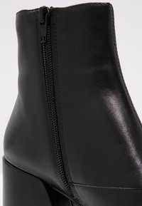 Bottine en cuir noir avec fermeture éclair latérale et talon bloc, présentant une texture lisse et des coutures détaillées.